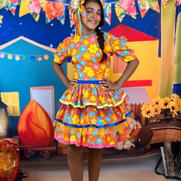 Menina com vestido junino laranja florido em frente ao painel de festa junina